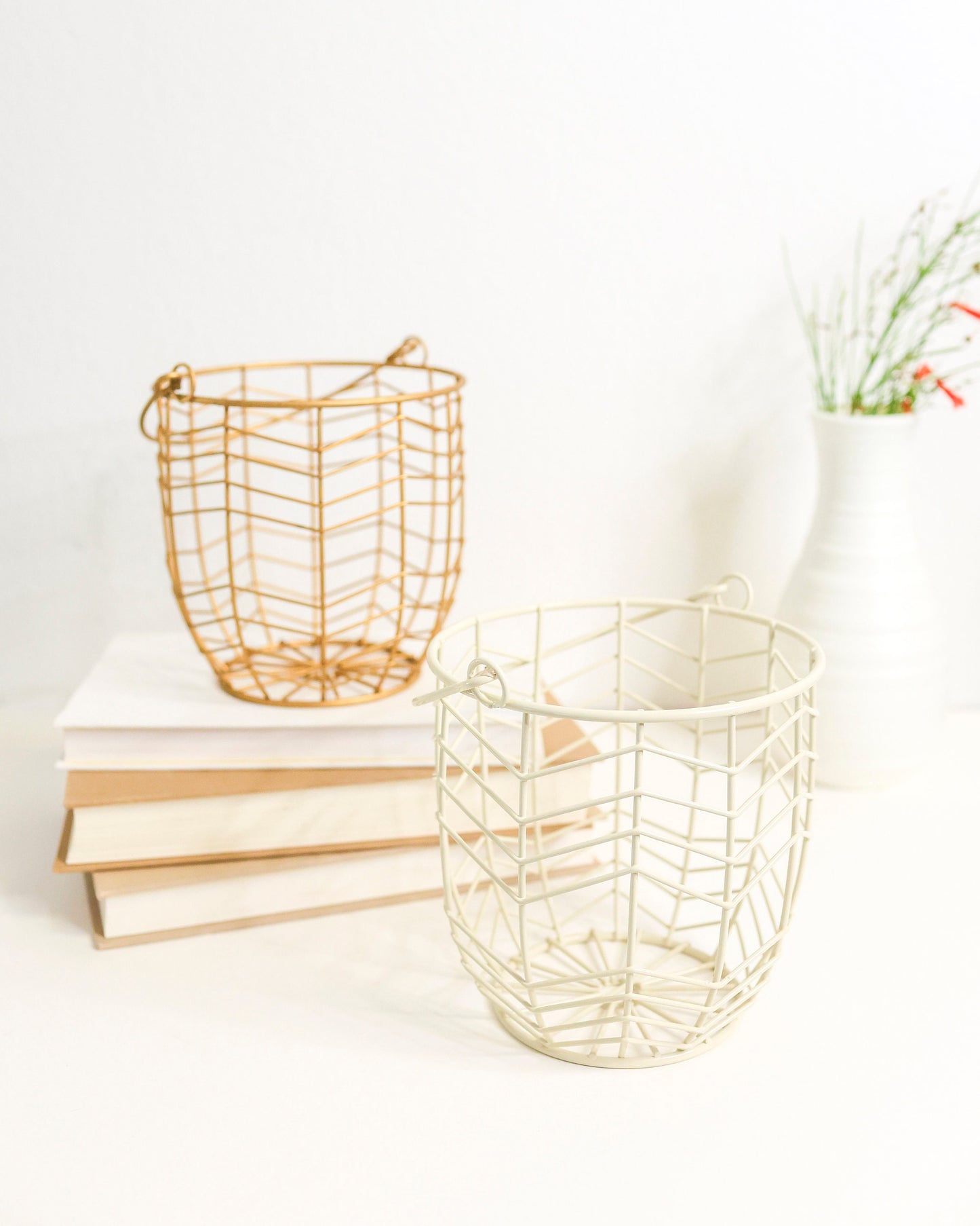 small metal gift basket with geometric wire pattern in gold or ivory. Shown on a white shelf with books and greenery out of focus behind.
