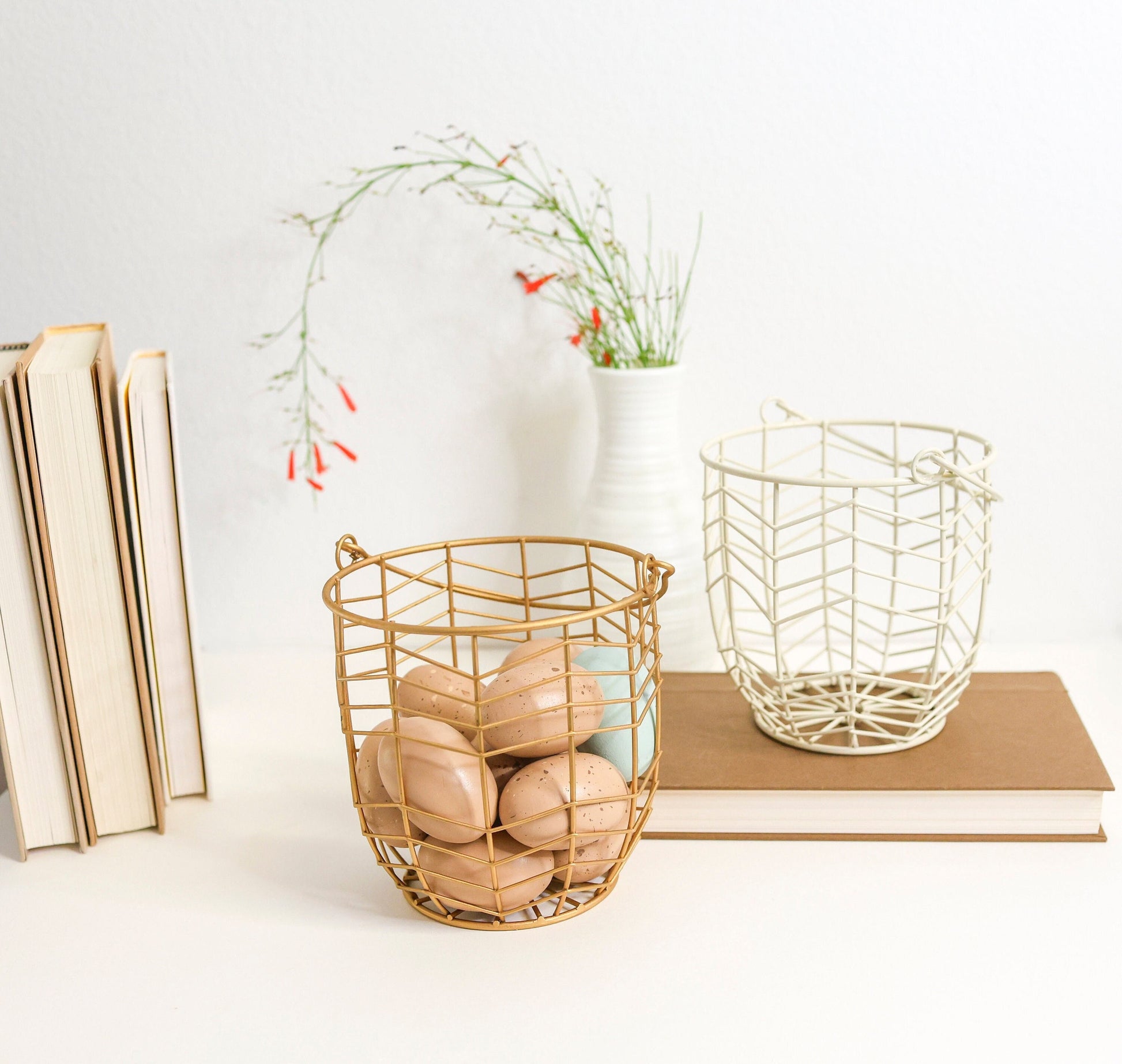 small metal gift basket with geometric wire pattern in gold or ivory. Shown on a white shelf with books and greenery out of focus behind.