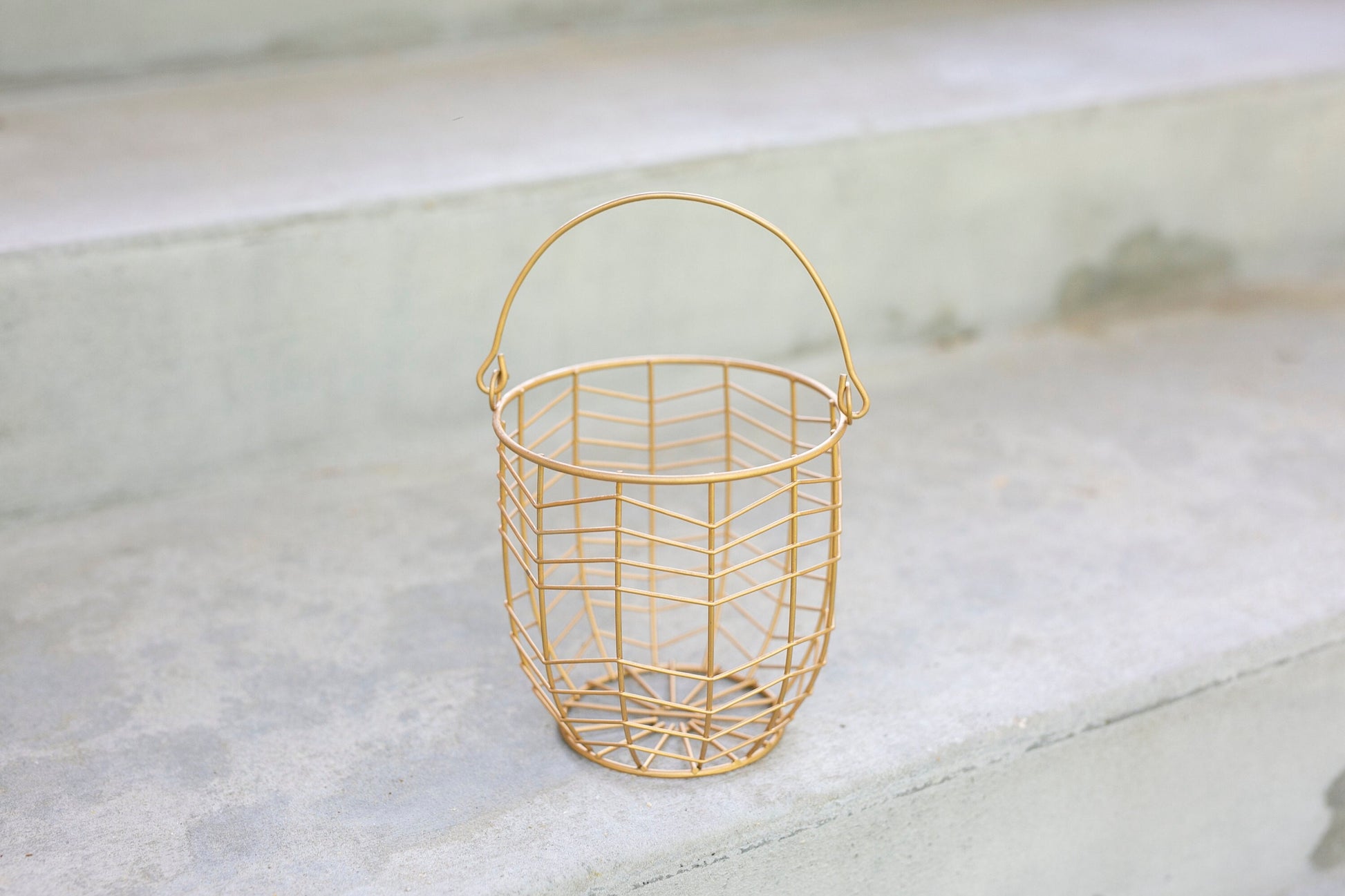 small metal gift basket with a geometric wire pattern in soft gold finish. shown on a soft concrete surface.