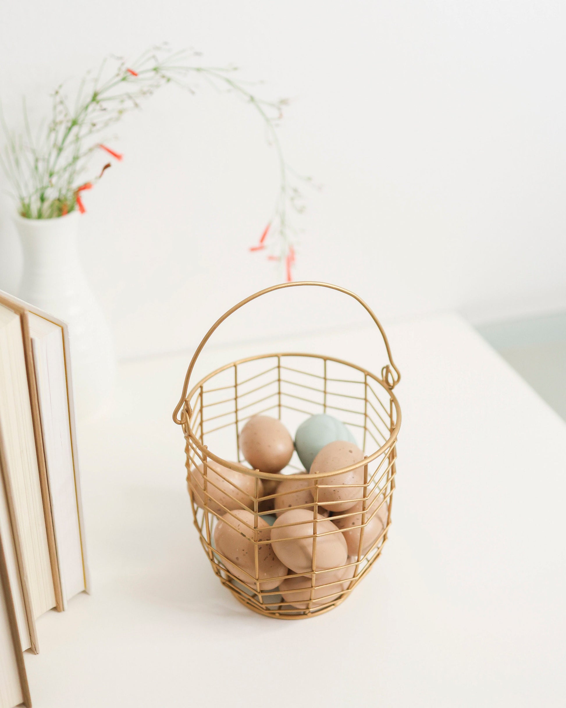 small metal gift basket with geometric wire pattern in gold. Shown on a white shelf with books and greenery out of focus behind.
