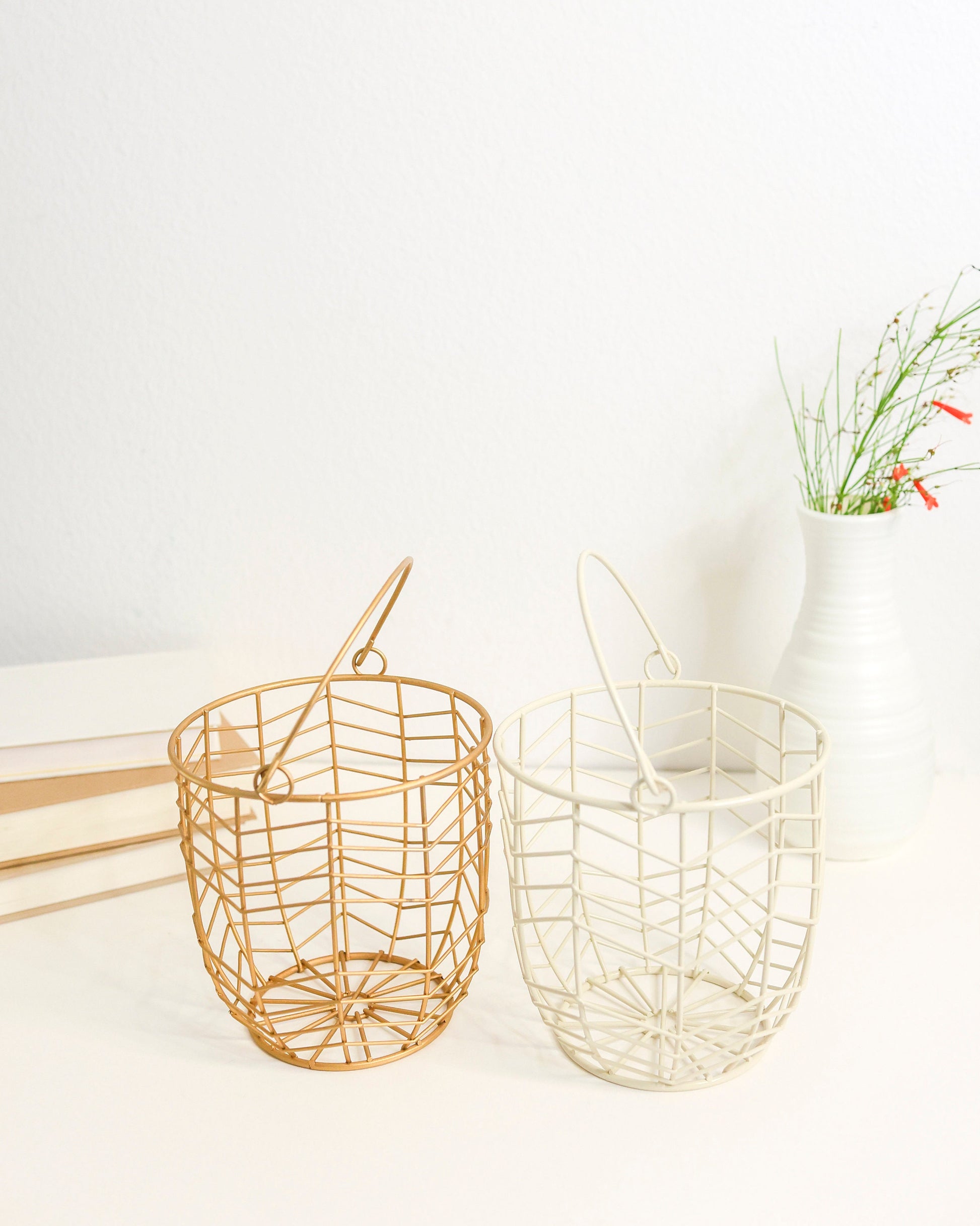 small metal gift basket with a geometric wire pattern in gold. Shown on a white shelf with books and greenery out of focus behind.